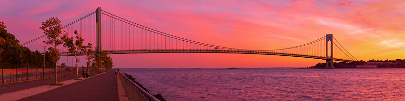 High resolution stitched panorama of the Verrazano-Narrows Bridge at Orange Yellow Blue Sunset. People are enjoying the view and taking pictures of the Majestic Sunset. The bridge connects boroughs of Brooklyn and Staten Island in New York City. The bridge was built in 1964 and is the largest suspension bridge in the USA. Historic Fort Wadsworth is under the bridge. The photo was taken from the Shore promenade in Bay Ridge Brooklyn. Canon EOS 6D full frame censor camera. Canon EF 85mm F/1.8 lens. 4:1 Image Aspect Ratio. This image was downsized to 50MP. Original image resolution was 80.6MP or 17956 x 4489px.