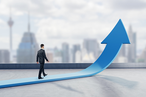 Young businessman walking on abstract upward blue arrow on blurry city background. Growth, career and growing finance concept