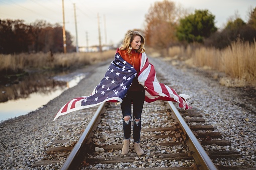 A blonde woman with the US flag standing on the railways surrounded by greenery under sunlight