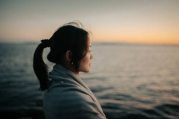 istock Asian woman cuddled with a blanket on the coastline looking aside during sunset 1440012859
