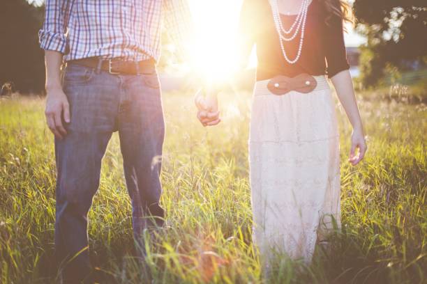 belle photo d’un couple se tenant la main debout dans un champ herbeux avec le soleil en arrière-plan - coulpe photos et images de collection