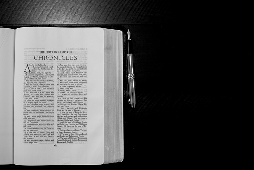 An overhead shot of an open bible near a fountain pen on a wooden surface in black and white