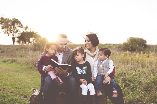 A beautiful cheerful family with a mother, father and three kids reading the Bible in the park