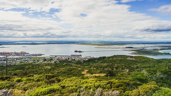 A panoramic view of a ship sailing in the sea near a green forest in Invercargill, New Zealand on a cloudy day