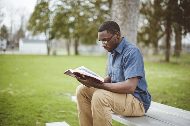 joven afroamericano sentado en el banco y leyendo la biblia - reading religious text black bible fotografías e imágenes de stock