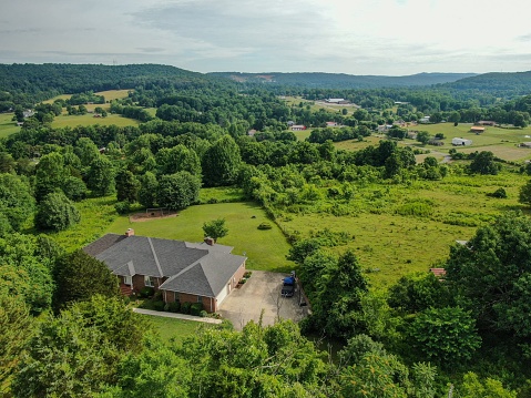 Cookeville, United States – July 06, 2022: An aerial view of a house on a green meadow in Cookeville, Tennessee,surrounded by trees,agricultural fields,and a forested mountain in the background