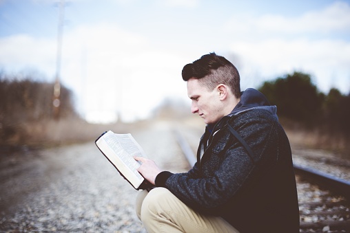 A shallow focus shot of a male sitting on train tracks and reading the bible