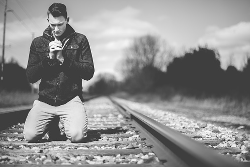 A grayscale shot of a male on his knees on the train tracks while praying