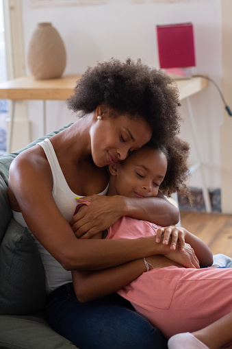 African American mother and daughter hugging tenderly. Woman and girl in casual clothes holding each other on coach. Love, family, bonding concept