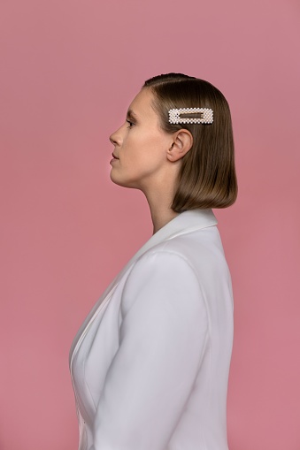 Profile view of young woman wearing white suit. Attractive female model is standing against pink background. She is wearing head accessory in studio.