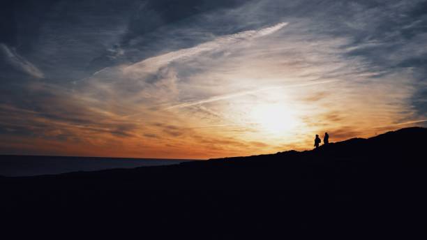 太陽を背景に丘の上を歩く2人のシルエットのワイドショット - walking night men sunset ストックフォトと画像