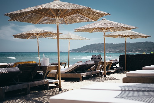 A selective wide shot of brown wooden loungers under parasols by the beach
