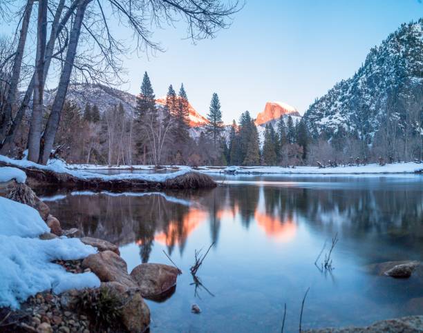 A wide landscape shot of a lake surrounded by rocks, trees and mountains over Half Dome in Yosemite during winter