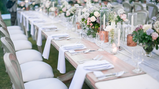 Festive outdoor wedding celebration table covered with natural tablecloth and beautifully and neatly served with sets of dishes, glasses, appliances and flower arrangements on a green background.