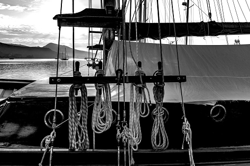 A greyscale closeup shot of ropes hanging in a sailing ship in the water under the cloudy sky