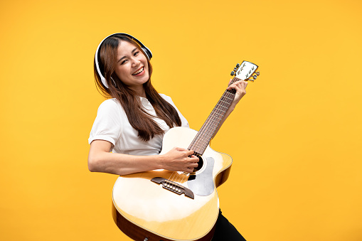 Young asian woman wearing headphone and white short sleeve shirt while playing guitar and singing rock song with happiness isolated over yellow background.