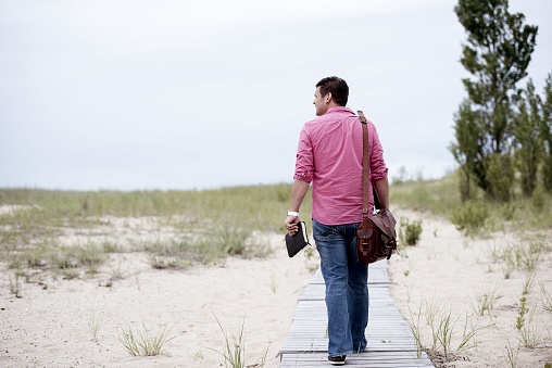 A male walking on a wooden pathway carrying his bag and holding the bible with a blurred background