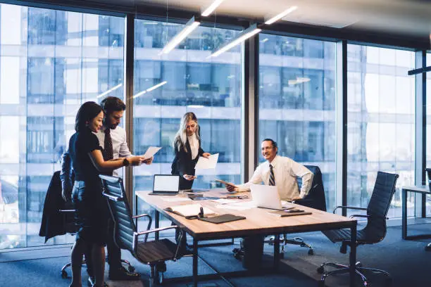 High angle of team of multiethnic business people in classy wear discussing business ideas and reading documents together while gathering at table with gadgets in modern office