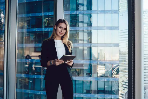 Young businesswoman in black suit standing at big window holding tablet and looking at camera smiling gently with modern building in background