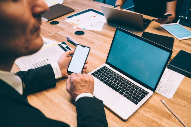 From above side view of crop male entrepreneur in formal suit sitting at table with laptop in contemporary workplace and browsing cellphone while working on project with colleagues