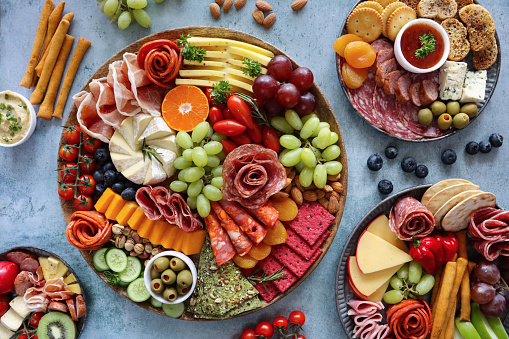 Stock photo showing close-up, elevated view of wooden charcuterie board covered with prepared sliced and chopped ingredients including rows of crackers, ham and salami roses, red and white grapes, grape tomatoes, pistachios, vine tomatoes, Brie, Red Leicester, Cheddar, almonds, blueberries, ramekin of stuffed olives.