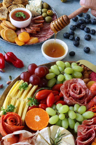 Image of unrecognisable person holding honey dipper over charcuterie boards, crackers, processed meat roses, grapes, cherry tomatoes, Brie, Red Leicester, Cheddar, nuts, blueberries, stuffed olives, mottled blue background, elevated view