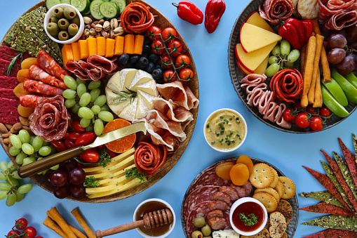 Stock photo showing close-up, elevated view of wooden charcuterie board covered with prepared sliced and chopped ingredients including rows of crackers, ham and salami roses, red and white grapes, grape tomatoes, pistachios, vine tomatoes, Brie, Red Leicester, Cheddar, almonds, blueberries, ramekin of stuffed olives.