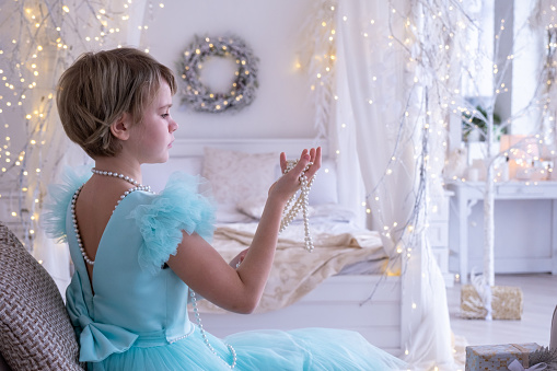 A beautiful teenage girl in a chic dress with an open back. A girl looks with interest at a Christmas present - a string of pearls. The interior of the bedroom is decorated for the new year and Christmas.