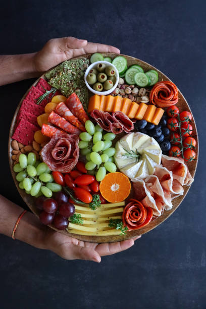 Stock photo showing close-up, elevated view of wooden charcuterie board covered with prepared sliced and chopped ingredients including rows of crackers, ham and salami roses, red and white grapes, grape tomatoes, pistachios, vine tomatoes, Brie, Red Leicester, Cheddar, almonds, blueberries, ramekin of stuffed olives. imagen de tablero de charcutería sostenido por una persona irreconocible, hileras de galletas, rosas de jamón y salami, uvas, pistachos, tomates de vid, brie, leicester rojo, cheddar, almendras, arándanos, ramekin de aceitunas rellenas, fondo negro, vis
