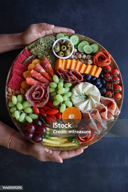 Image Of Charcuterie Board Held By Unrecognisable Person Rows Of Crackers Ham And Salami Roses Grapes Pistachios Vine Tomatoes Brie Red Leicester Cheddar Almonds Blueberries Ramekin Of Stuffed Olives Black Background Elevated View Stock Photo - Download Image Now