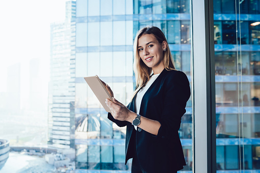 Half length portrait of cheerful female intelligent manager looking at camera using digital tablet for research in office, smiling business woman in elegant wear banking on touchpad enjoying gadget