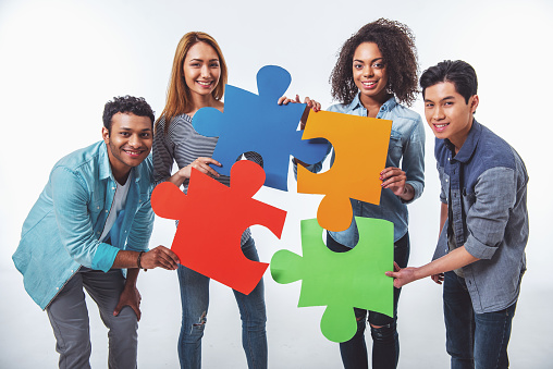 Young people of different nationalities with colorful pieces of puzzle are looking at camera and smiling, isolated on white background