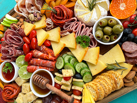 Stock photo showing close-up, elevated view of wooden charcuterie board covered with prepared sliced and chopped ingredients including rows of crackers, ham and salami roses, sliced cucumber, apple, dried apricots, cherry tomatoes, Red Leicester, pistachios, blueberries, almonds, orange and limes.