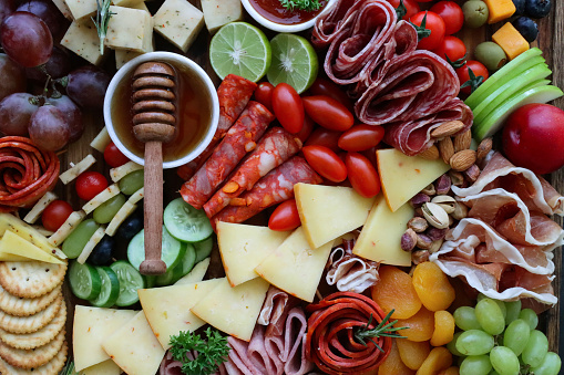 Stock photo showing close-up, elevated view of wooden charcuterie board covered with prepared sliced and chopped ingredients including apricots, crackers, ramekin of honey, salami roses, red and white grapes, cherry vine tomatoes, apple slices, cheddar and herb cheese.