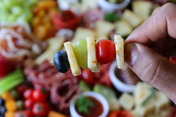 Stock photo showing elevated view of wooden charcuterie board covered with prepared sliced and chopped ingredients including bread sticks, beetroot crackers, ramekin of chutney, salami roses, red grapes, cherry vine tomatoes, kiwi half, cheddar and herb cheese cubes. imagen en primer plano de una persona irreconocible sosteniendo kebab de queso y palillo de cóctel de frutas sobre tabla de charcutería, galletas, ramekin de chutney, rosas de salami, uvas rojas, tomates cherry vine, mitad de kiwi, cubos de queso cheddar
