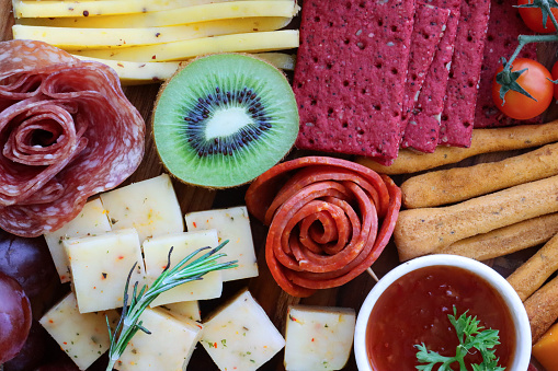 Stock photo showing close-up, elevated view of wooden charcuterie board covered with prepared sliced and chopped ingredients including bread sticks, beetroot crackers, ramekin of chutney, salami roses, red grapes, cherry vine tomatoes, kiwi half, cheddar and herb cheese cubes.