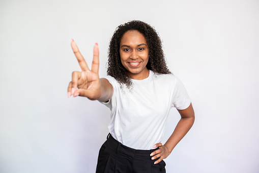 Portrait of happy young woman making victory gesture. African American lady wearing white T-shirt looking at camera and smiling over white background. Happiness concept