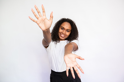 Portrait of happy young woman stretching arms for embrace over white background. African American lady wearing white T-shirt showing palms and smiling. Happiness concept