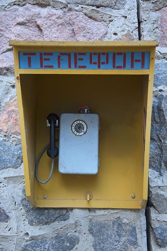 Close-up view of vintage street phone in yellow booth hangs on a gray brick wall. Text on the top “Телефон” in English Phone.