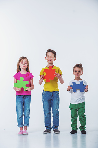 Full length portrait of cute little kids in casual clothes holding colorful puzzles, looking at camera and smiling, isolated on a white background