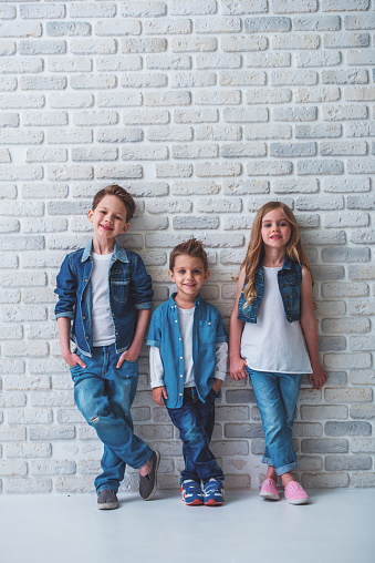 Full length portrait of cute little kids in stylish jeans clothes looking at camera and smiling, standing against white brick wall