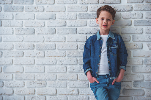 Stylish little boy in jeans clothes is looking at camera and smiling, standing with hands in pockets against white brick wall