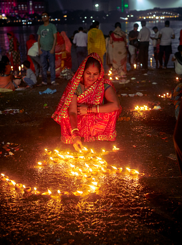 Kolkata, 11/07/2022: People in beautiful ethnic dresses, illuminating ghats of Ganges river by decorating with diyas (handmade, earthen oil lamps), on the day of Dev Deepawali (or Diwali).\nDev Deepawali is an age old traditional religious festival mostly celebrated on Kartik Poornima, the full moon of the Hindu month of Kartik, fifteen days after Diwali.