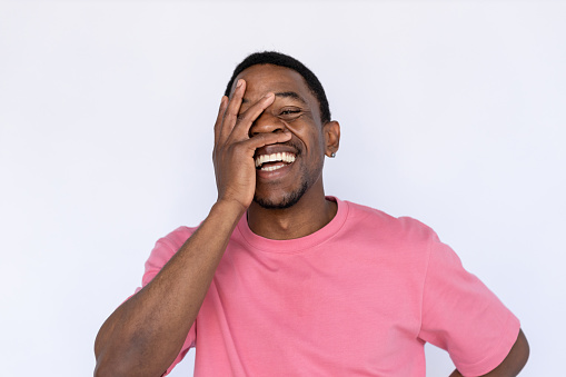 Portrait of amused African American man laughing. Happy young male model with short dark hair in pink T-shirt looking at camera, smiling, covering face with hand. Humor, happiness concept
