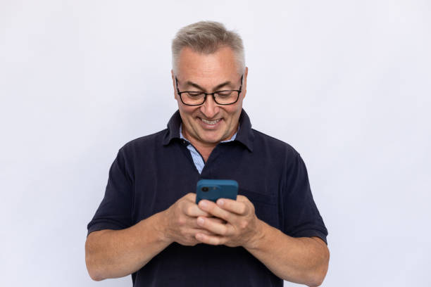 Senior man typing on phone Senior man typing on phone. Male model in blue polo shirt messaging, texting on smartphone with smile. Portrait, studio shot, texting concept one senior man only stock pictures, royalty-free photos & images