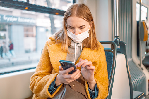 Young caucasian woman wear protective face mask, using mobile smart phone, chatting in social media, sitting in public bus during pandemic Covid-19. New normal concept.