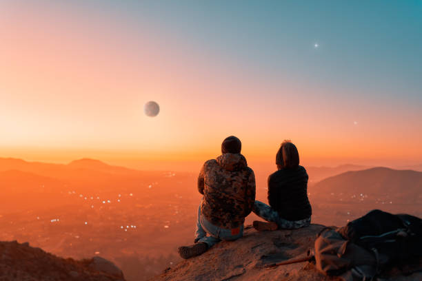 silhouette of a couple sitting on top of a hill enjoying the sunset, back view. stock photo