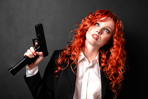 Portrait of a young red-haired woman armed with a pistol in a business suit. The concept of the business mafia.