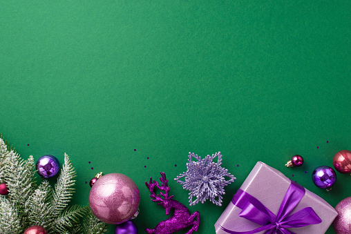 Christmas Day concept. Top view photo of lilac giftbox with purple ribbon bow pink violet baubles reindeer flower ornaments fir branches in snow confetti on isolated green background with blank space