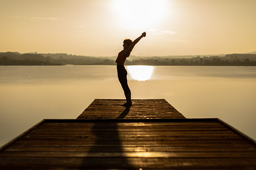 Mature woman doing yoga on wooden pier above the beautiful lake at sunset. She is dedicated to healthy lifestyle